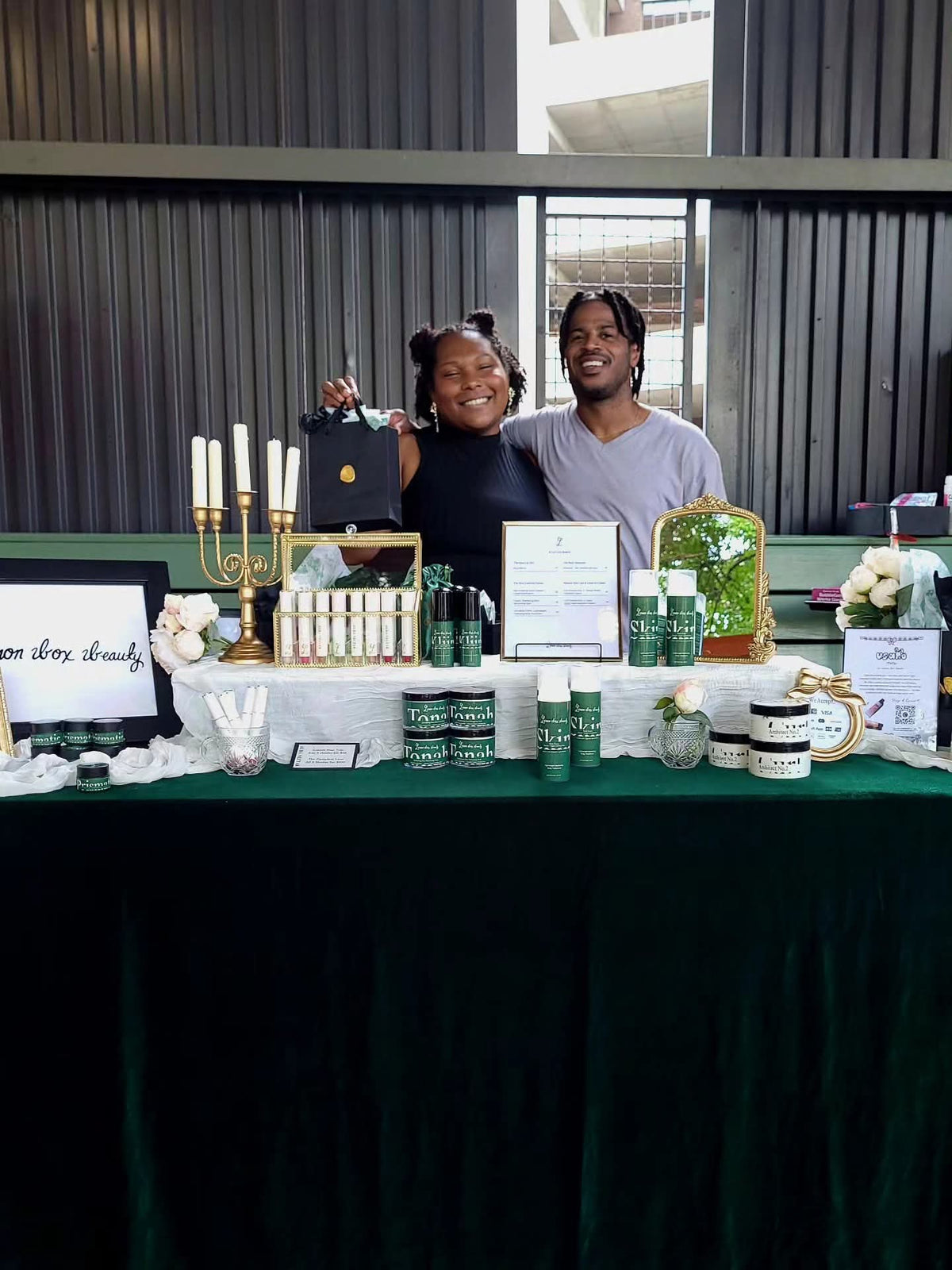 A smiling couple stands behind a decorated product display table at what appears to be an outdoor market or event. The table is covered with a dark green cloth and displays multiple skincare or beauty products, including green-and-white bottles, jars, a candelabra, and branded signs.