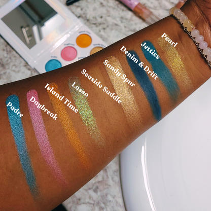 Color swatches on a person's arm with labeled shades, including 'Daybreak' and 'Padre'. Makeup palette with various colors held by a hand, Seaside Lasso Eyeshadow palette