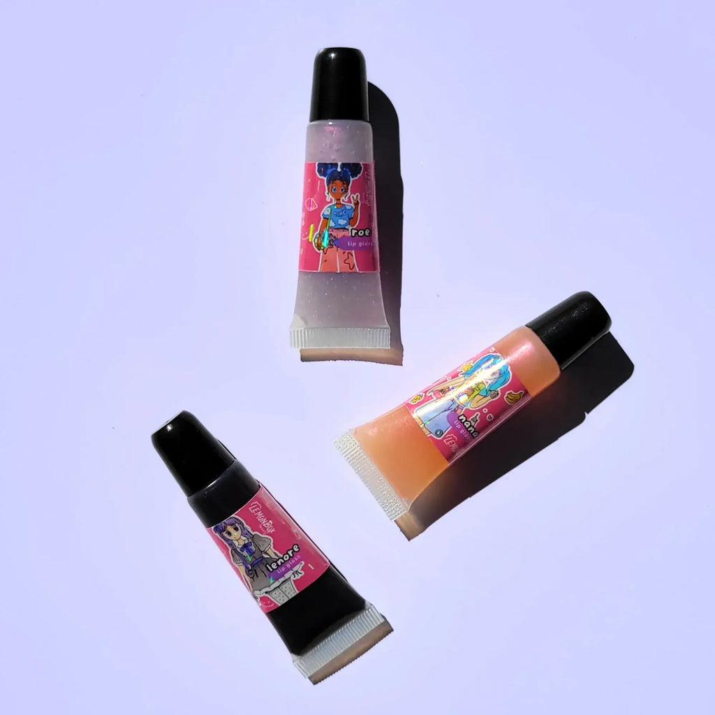 A trio of Lemon Box Beauty Anime Shine Lip Glosses—shades Roe, Nana, and Lenore—translucent iridescent, peachy-pink, and deep plum-purple vegan lip oils in squeeze tubes on a lavender background.