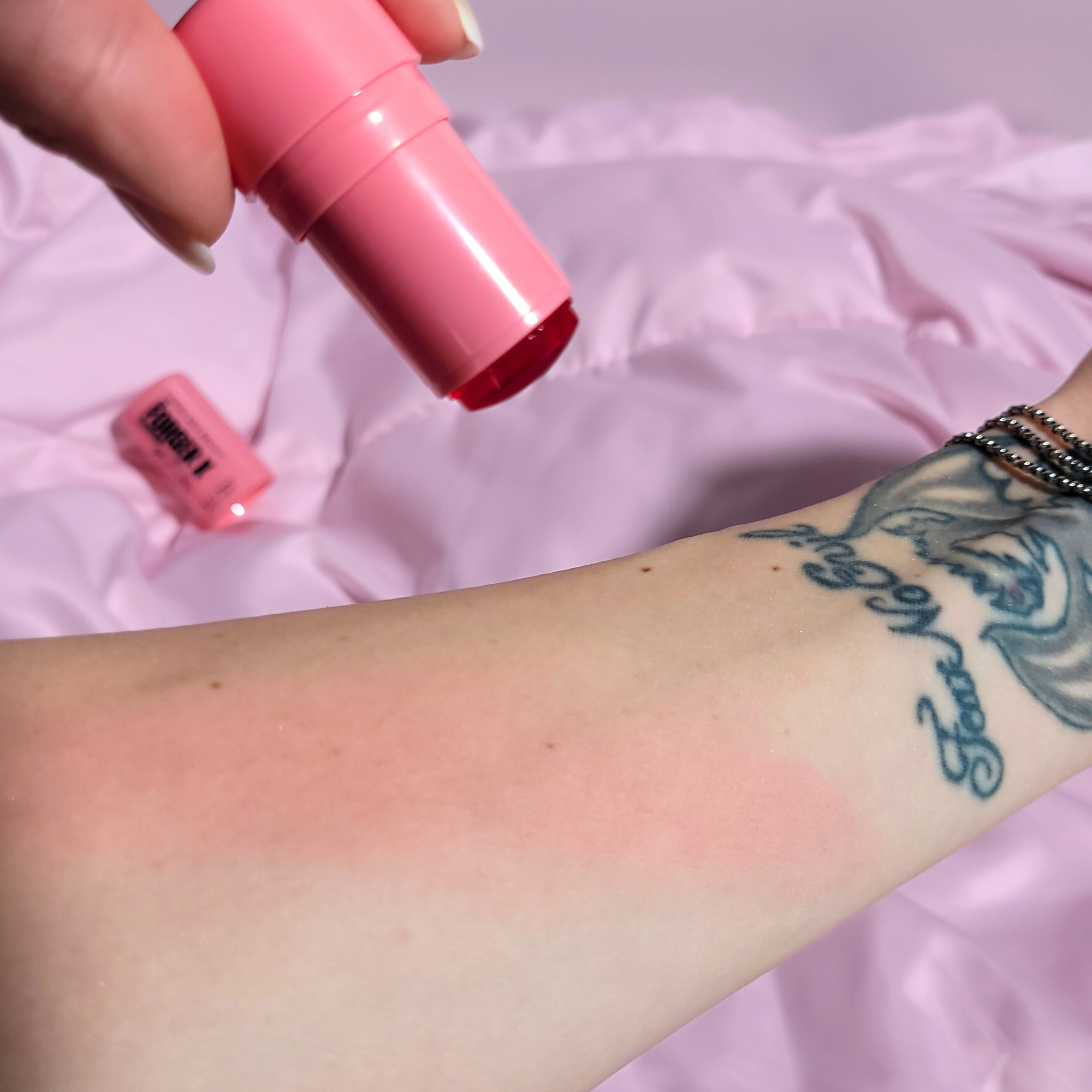 Pink jelly cheek tint being applied to a person's arm with a tattoo on a pink background