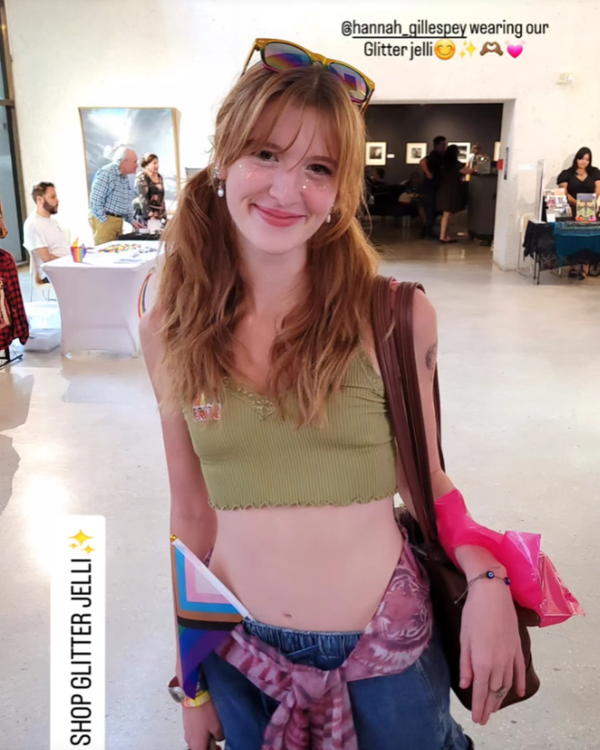 Woman in a green crop top and denim jacket standing in a room with people and tables wearing glitter jelly makeup on face