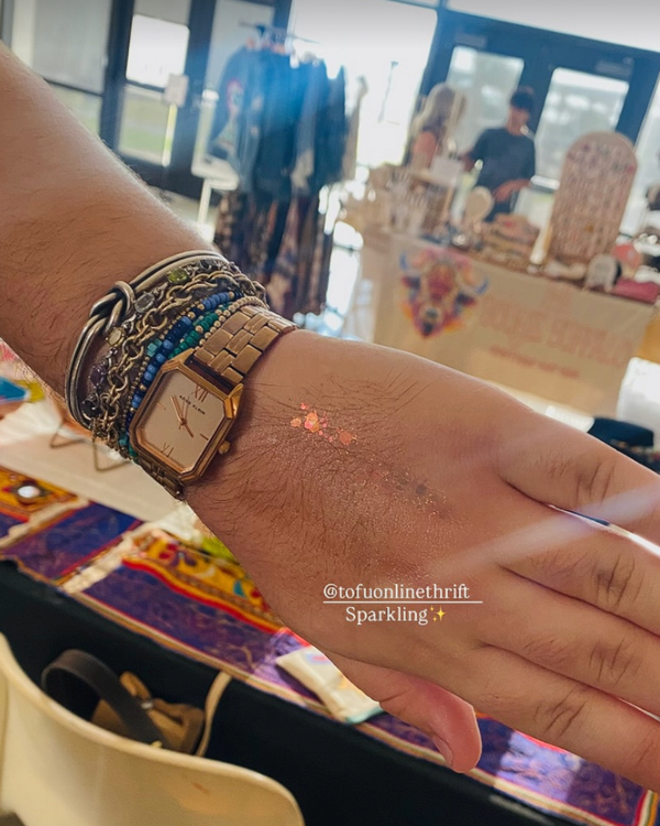 Hand wearing a gold watch with multiple bracelets in a blurred indoor wearing glitter jelly makeup on hand