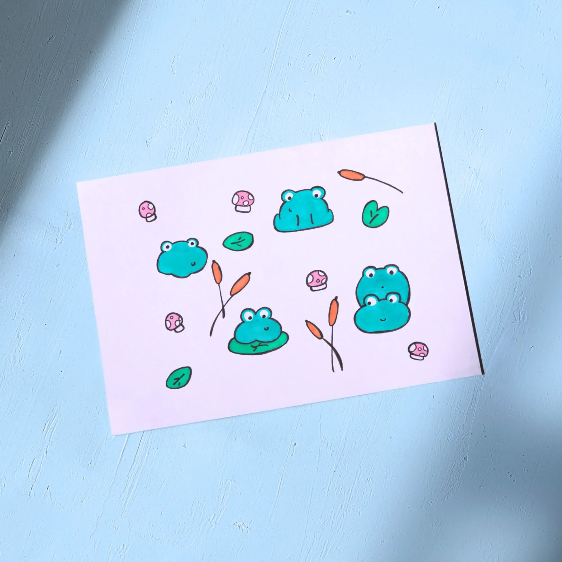 White Greeting Card with frogs and mushrooms on a light blue background
