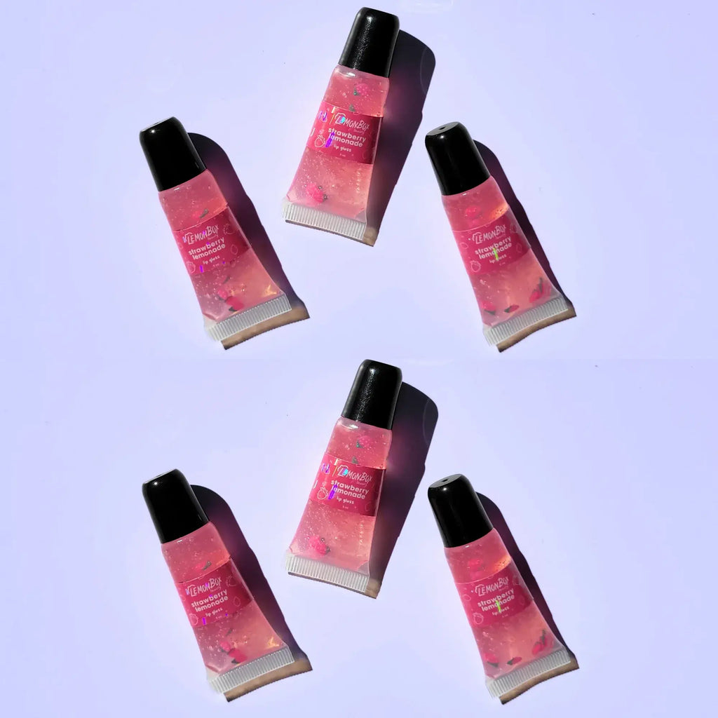 A trio of Lemon Box Beauty Strawberry Lemonade High-Shine Squeeze Lip Glosses—clear pink vegan lip oil tubes containing cute pink strawberry clay slices on a lavender background