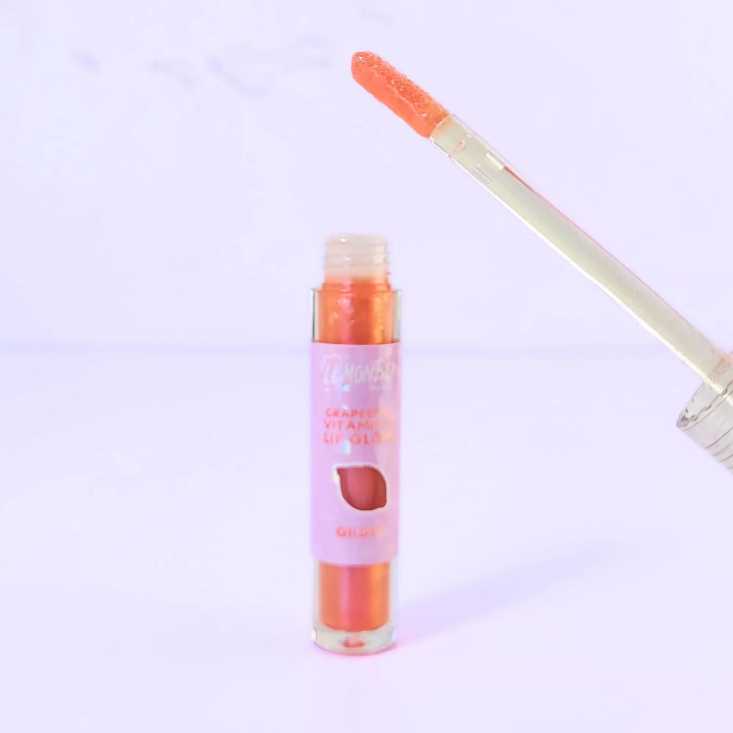 Lemon Box Beauty Grapeseed Vitamin E Lip Gloss in shade Gilded - shimmering copper-gold hydrating lip oil in a clear tube against a lavender background.