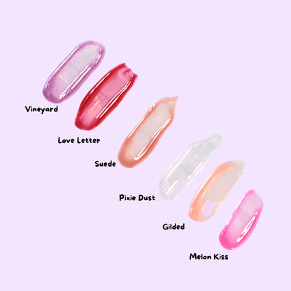 Lemon Box Beauty collection of six lip gloss swatches on skin—shades Gilded, Love Letter, Melon Kiss, Pixie Dust, Suede, and Vineyard—showing shimmering and creamy finishes.