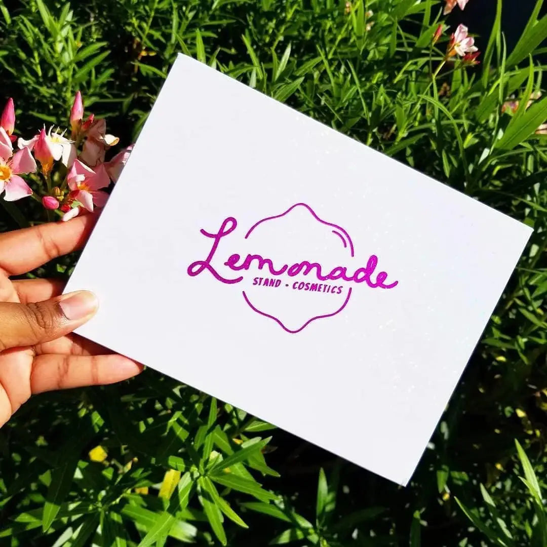 Hand holding a white card with 'Lemonade Stand Cosmetics' logo against a green outdoor background.