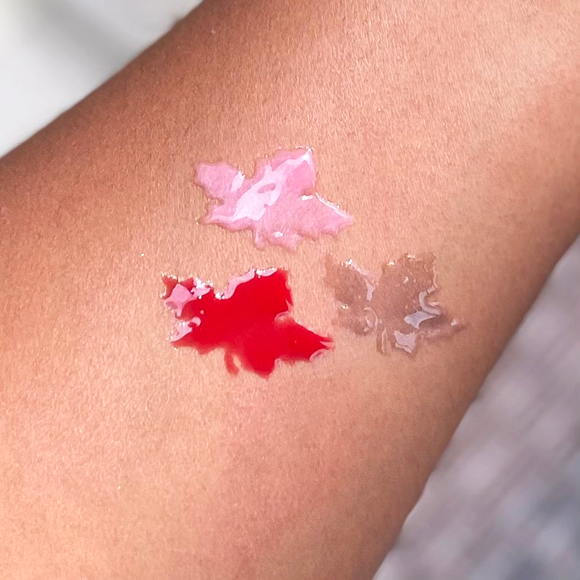Three droplets of different colored lip gloss swatches on a person's arm.