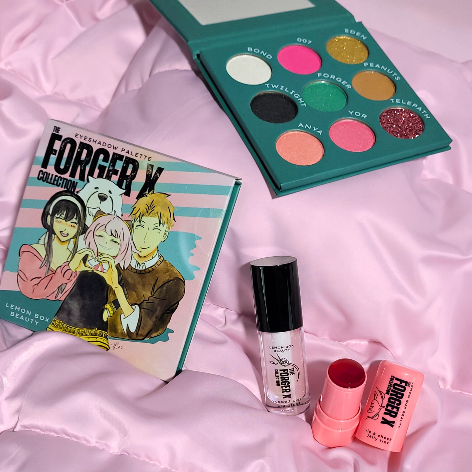 Makeup products including a palette, lip gloss, and lipstick on a pink fabric background