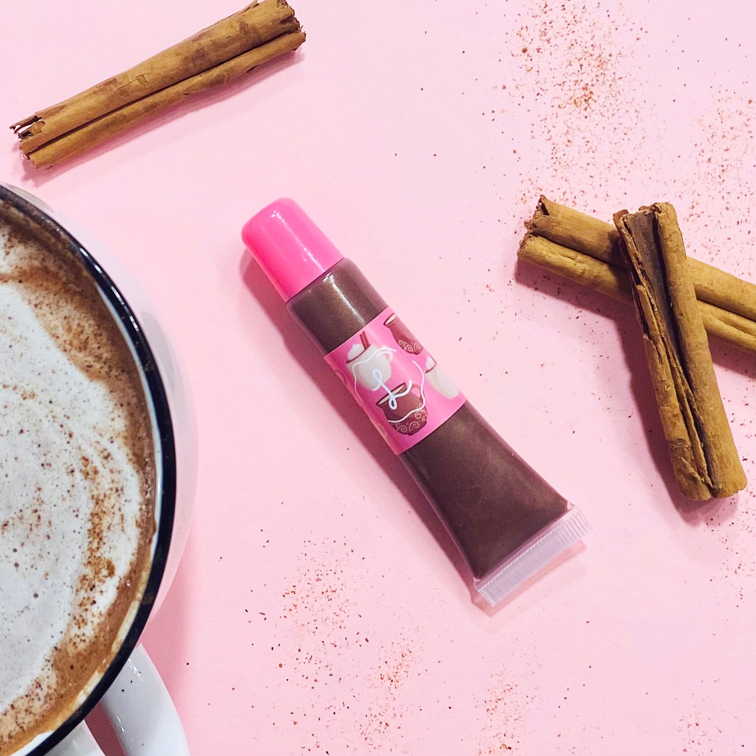 Chocolate lip balm with pink cap on a pink background with cinnamon sticks and a cup of coffee.