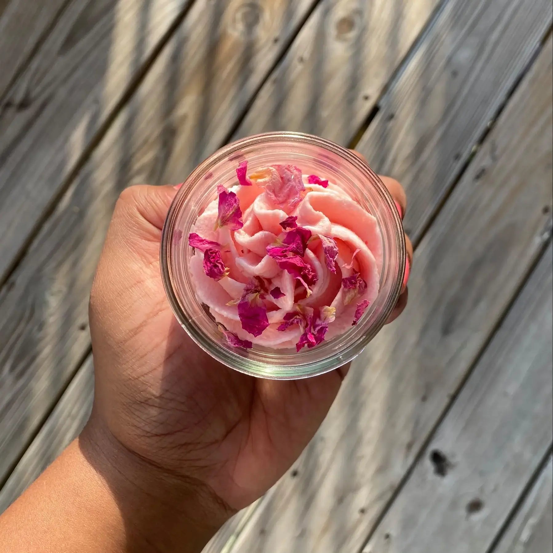 Hand holding a small container of pink dessert with rose petals against a wooden background