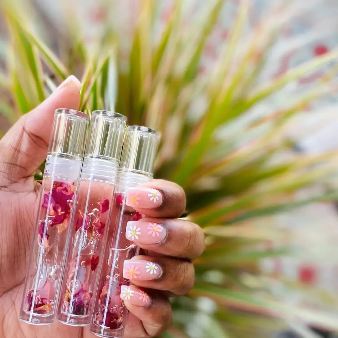 Hand holding three small transparent Rose lip gloss with floral elements against a blurred natural background