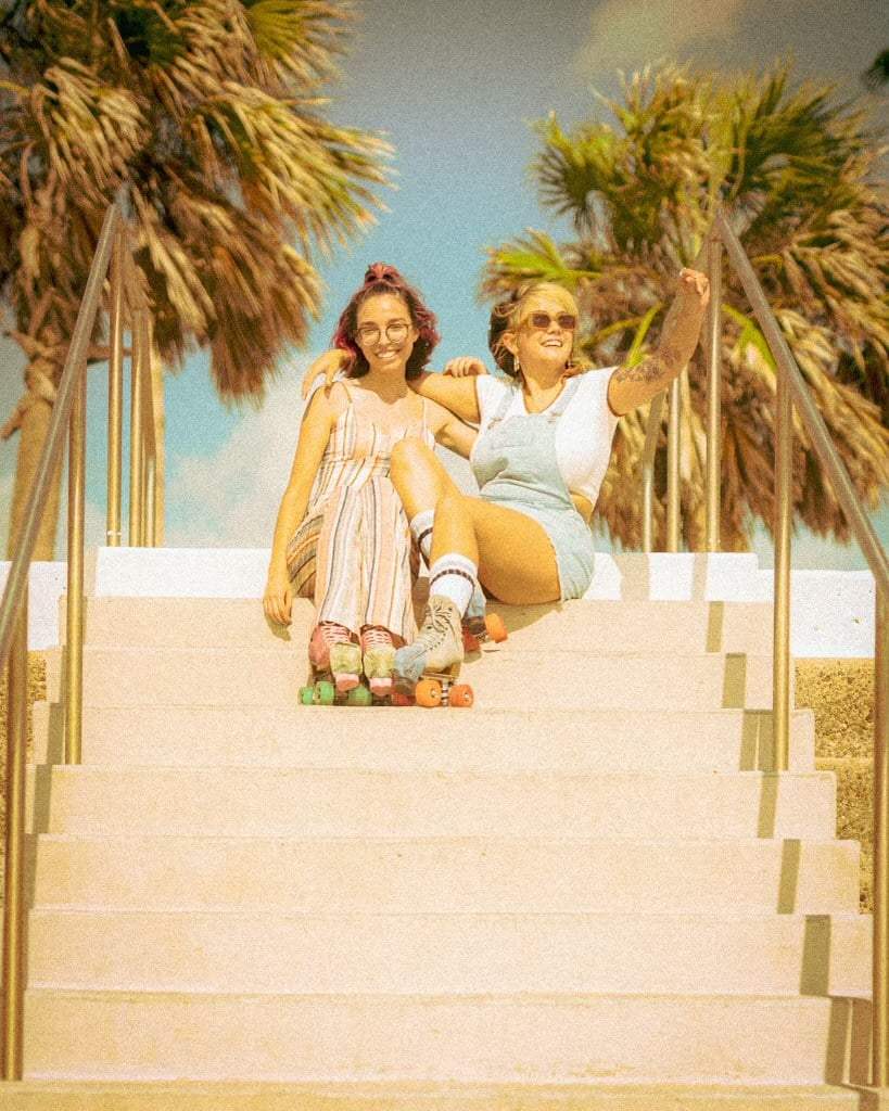 Two women sitting on a staircase with palm trees in the background Roller skates