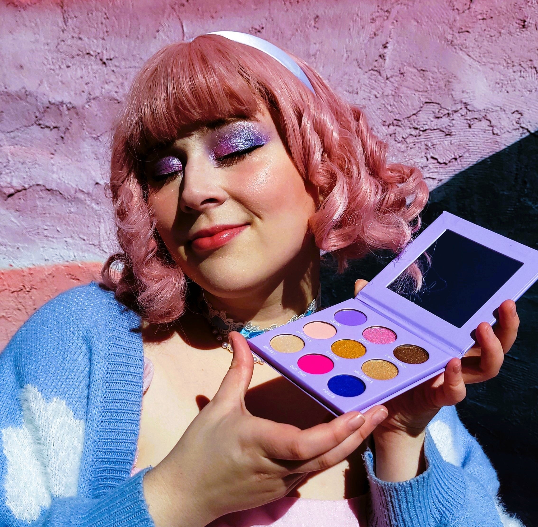 Person with pink hair holding a makeup palette against a textured pink wall.