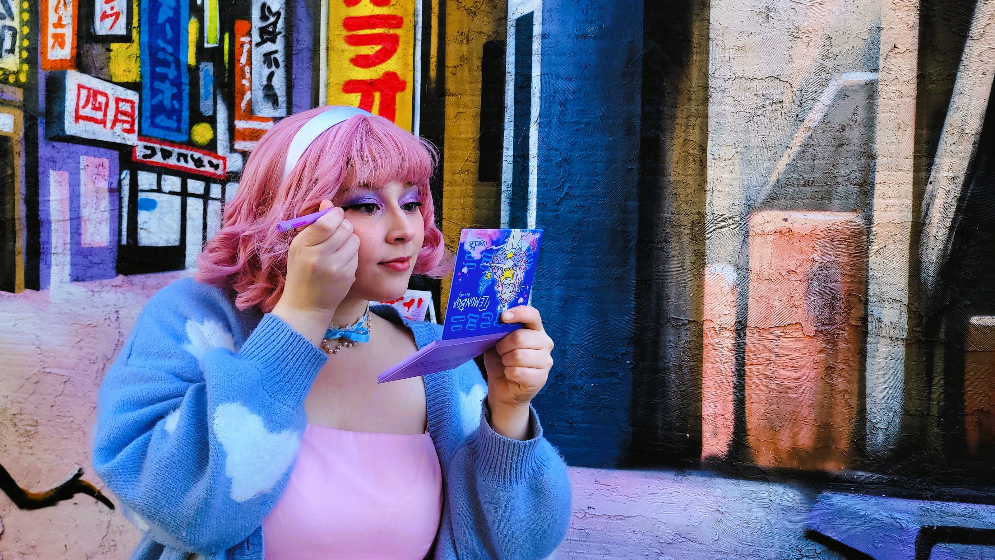 Person with pink hair and a blue sweater holding a colorful book in front of a graffiti-covered wall.