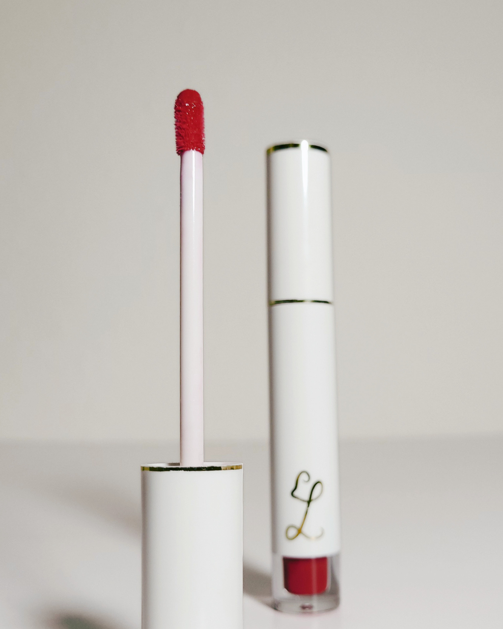 A white cosmetic tube with gold trim and a red lip color, shown with the applicator wand removed and standing in the cap. The brand logo is a stylized gold 'L' on the tube.