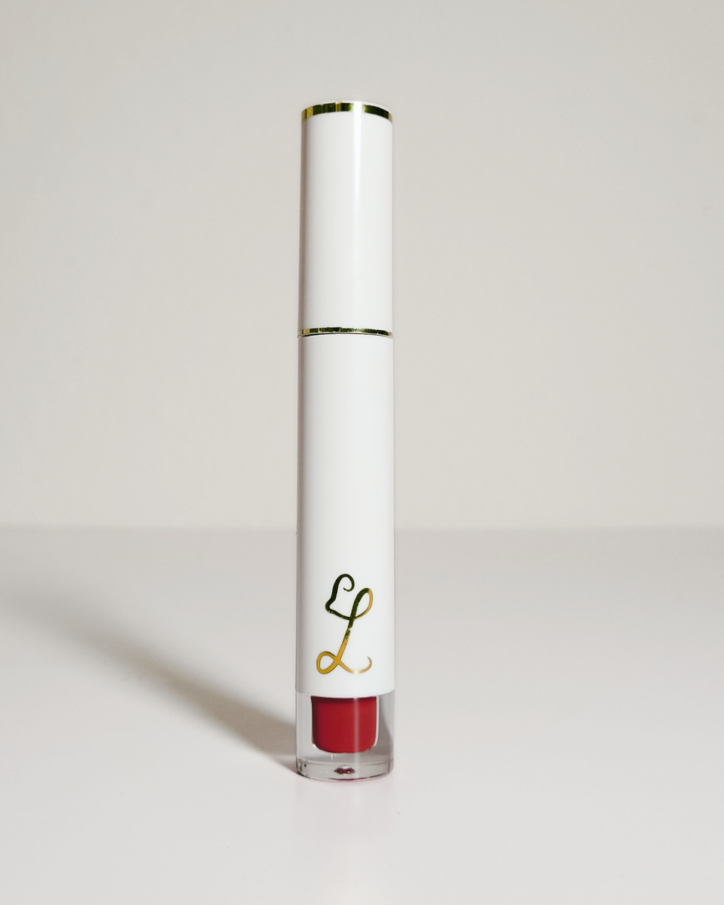 A white cylindrical liquid lipstick tube with gold trim and a red lip color visible at the bottom, standing upright on a light gray background.