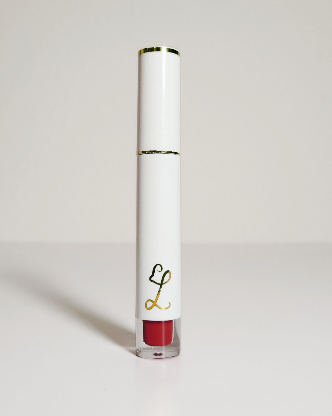 A white cylindrical liquid lipstick tube with gold trim and a red lip color visible at the bottom, standing upright on a light gray background.