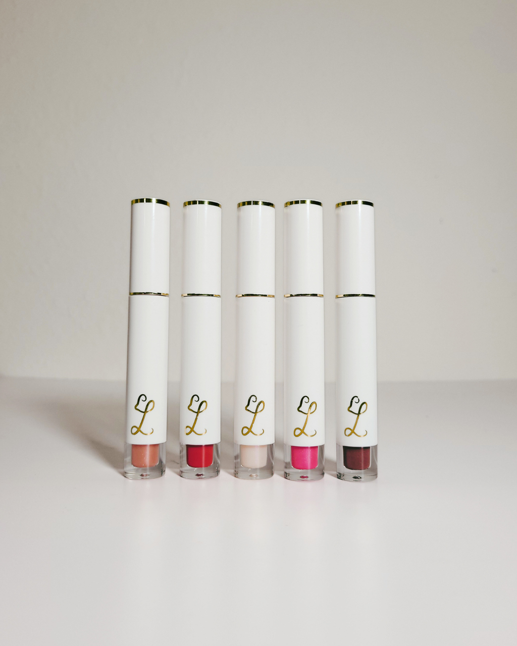 Five white lip gloss tubes with gold trim and a gold script 'L' logo, standing upright in a row on a light gray surface against a light gray background.