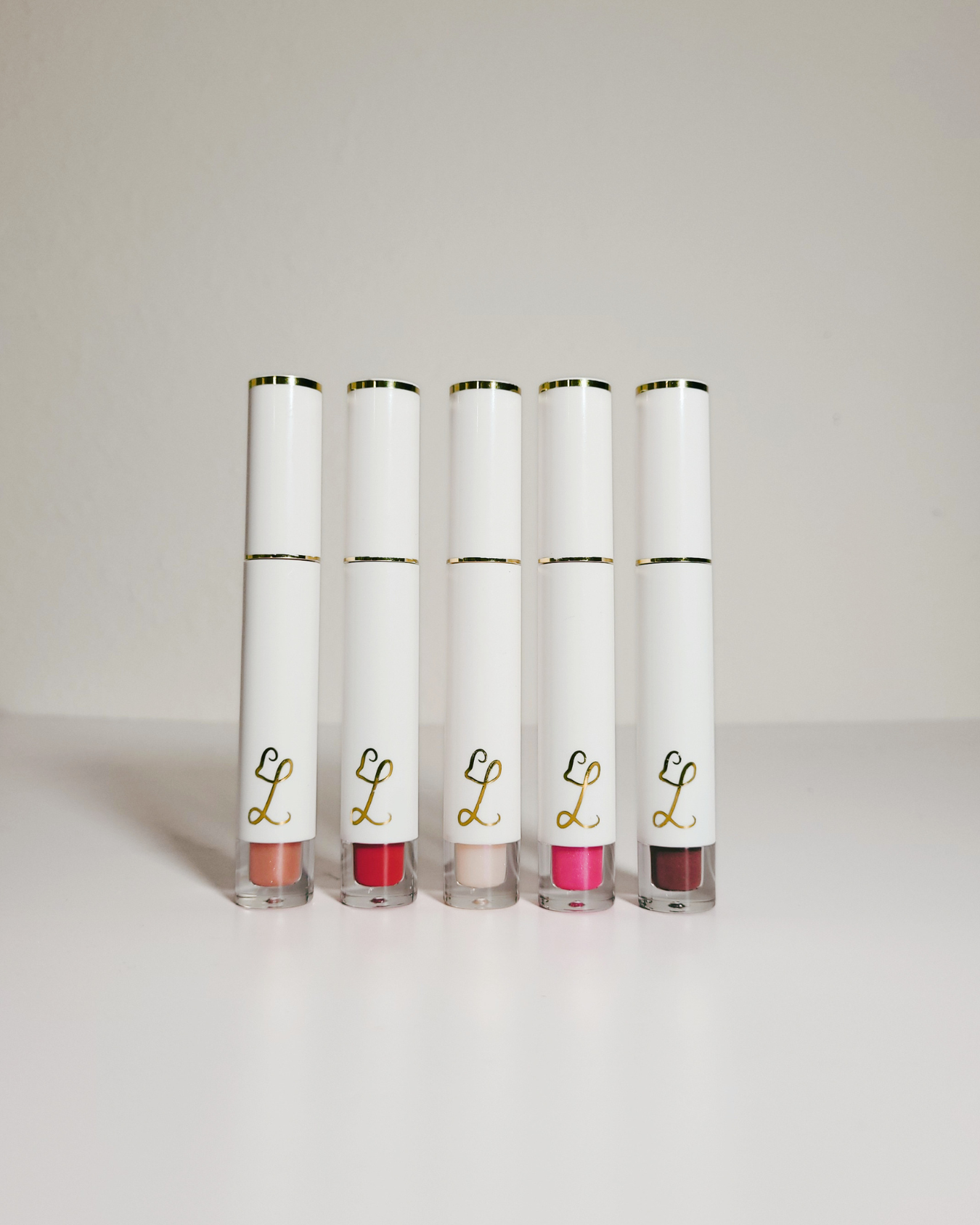 Five white lip gloss tubes with gold trim and a gold script 'L' logo, standing upright in a row on a light gray surface against a light gray background.