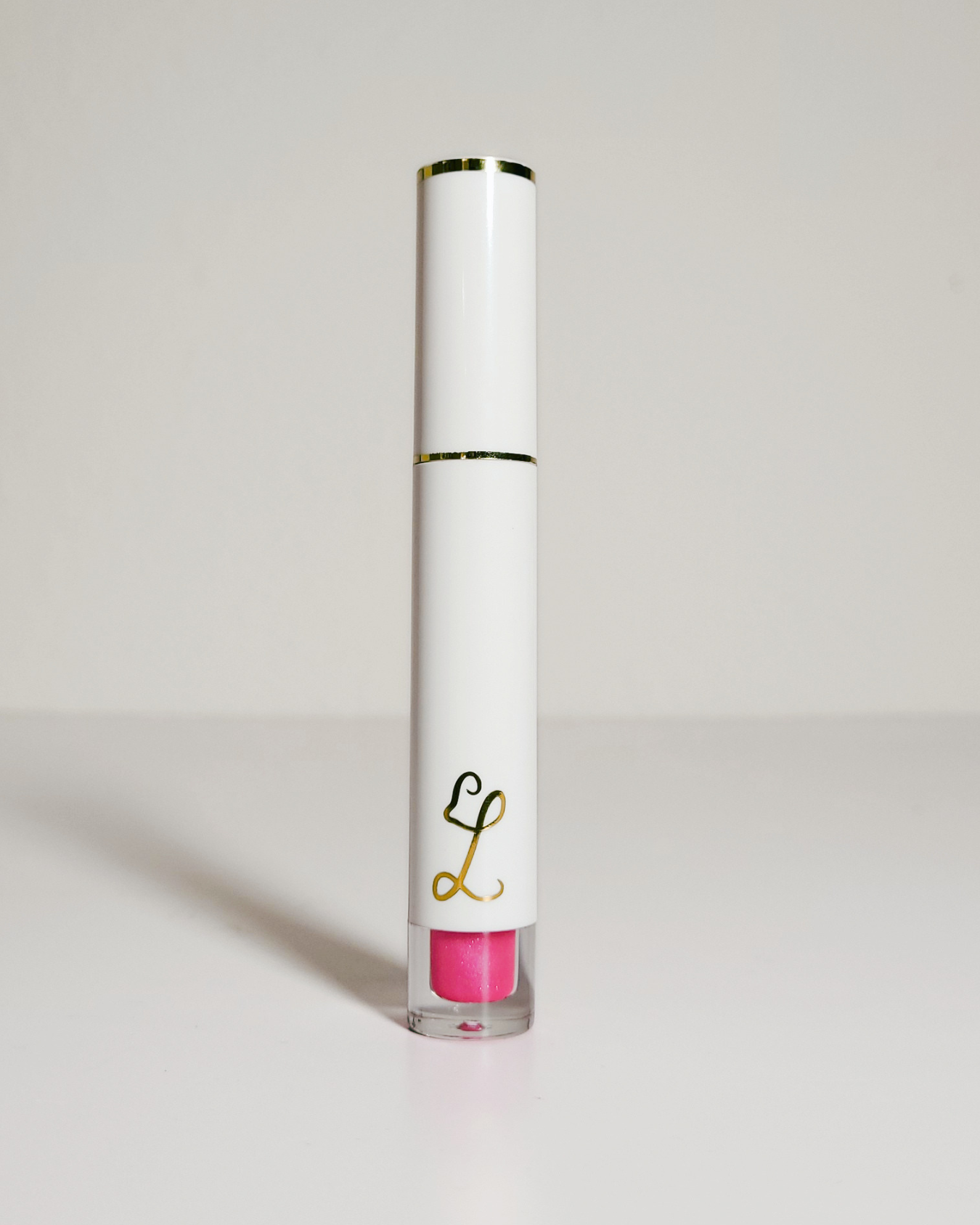 A white cosmetic tube with gold trim and a gold monogram logo, standing upright on a light gray surface against a light gray background. The clear lower section reveals a bright pink product inside.