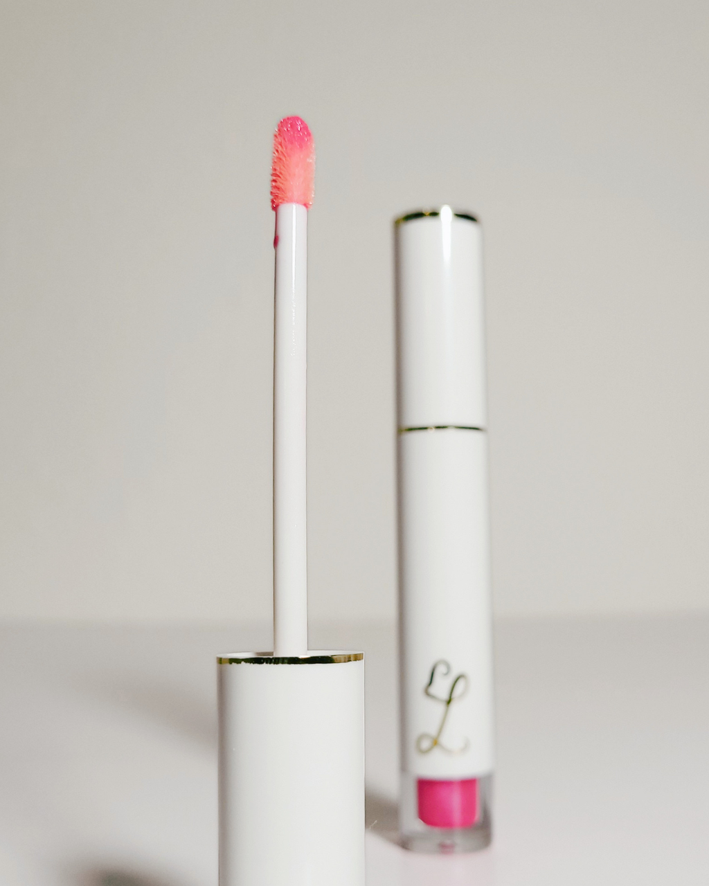 A white cosmetic tube with gold trim and a pink lip gloss applicator wand, shown next to the closed tube on a light gray background.