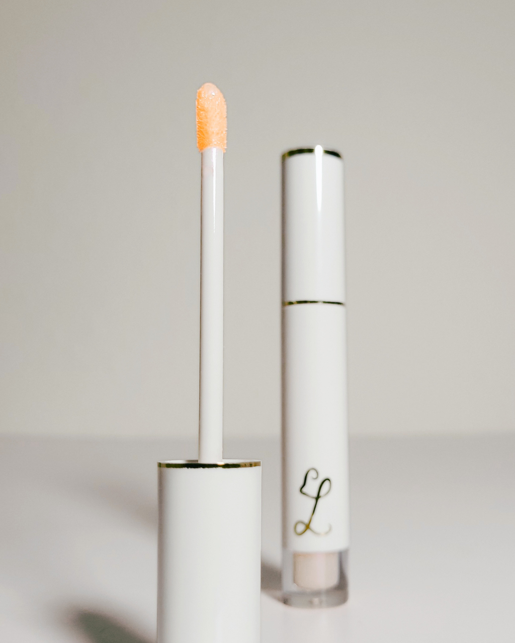 A white cosmetic tube with gold trim and a doe-foot applicator showing a peachy orange cream product, alongside the closed tube on a light gray background.