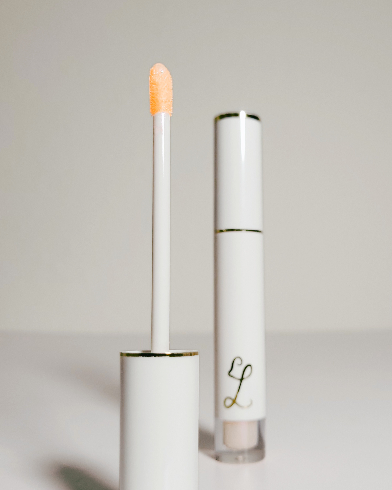 A white cosmetic tube with gold trim and a doe-foot applicator showing a peachy orange cream product, alongside the closed tube on a light gray background.