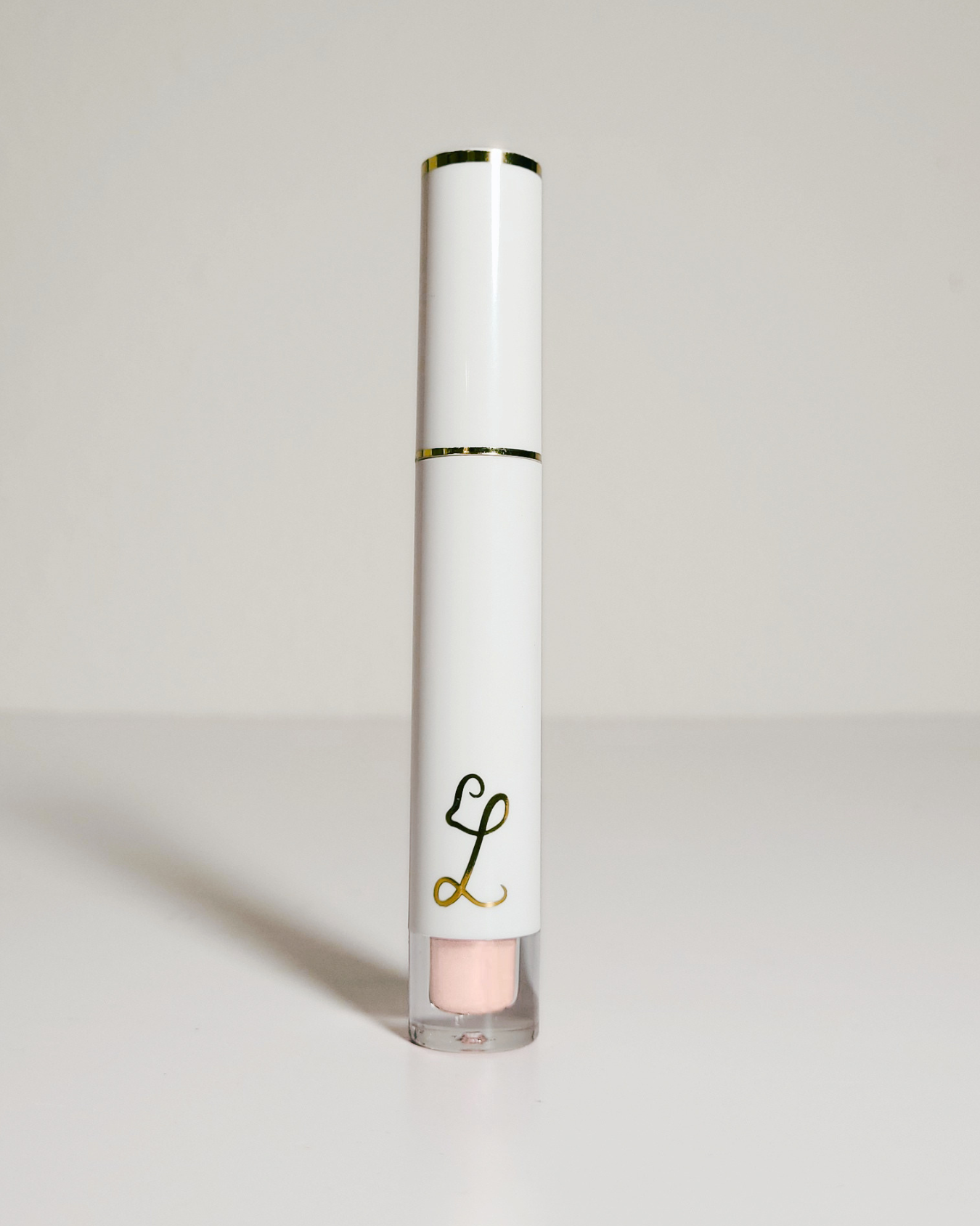 A white cosmetic tube with gold trim and a gold monogram logo, standing upright on a light gray surface against a light gray background.