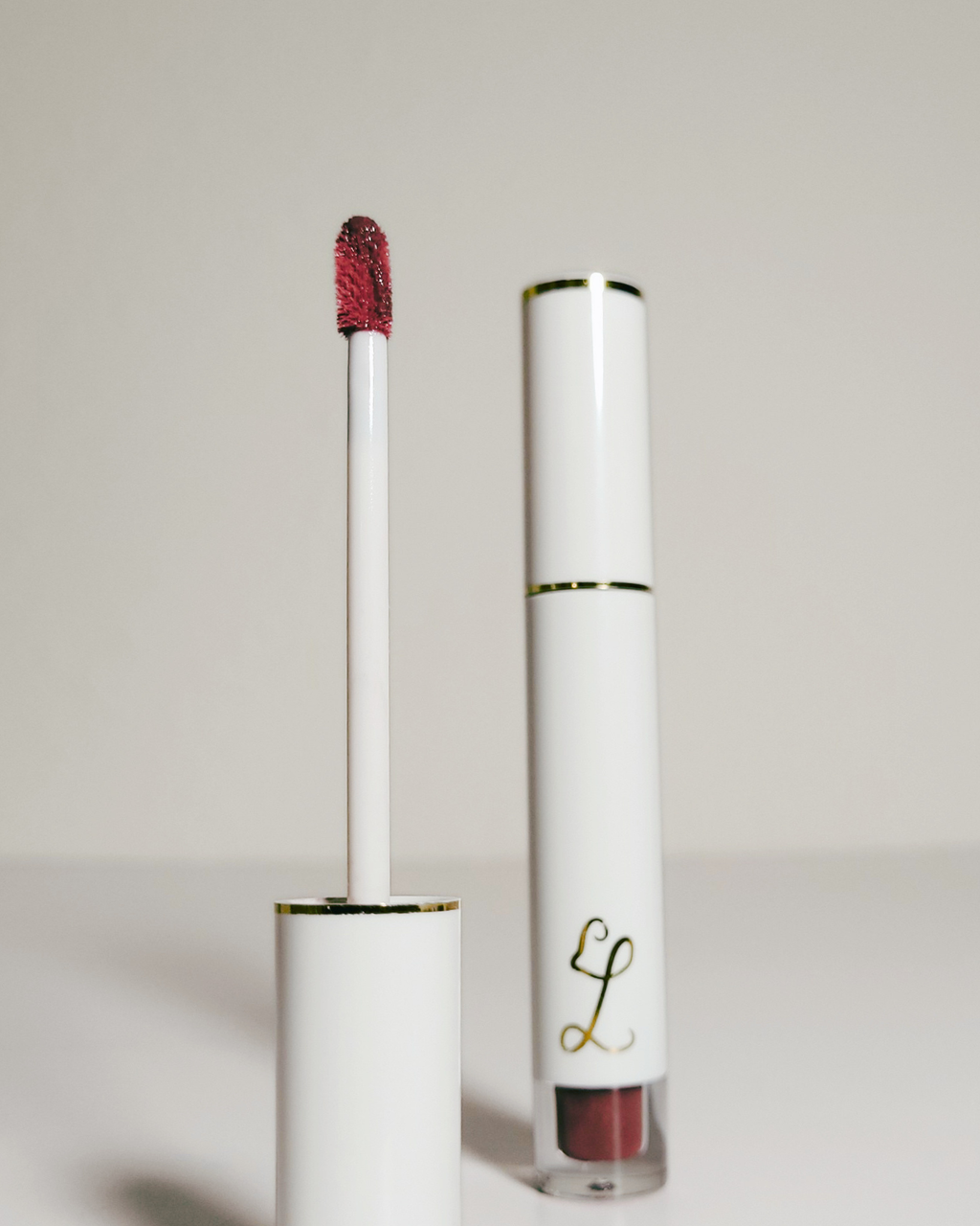 A white liquid lipstick tube with gold trim stands next to its applicator wand, which is loaded with a deep berry red shade, on a clean light gray background.