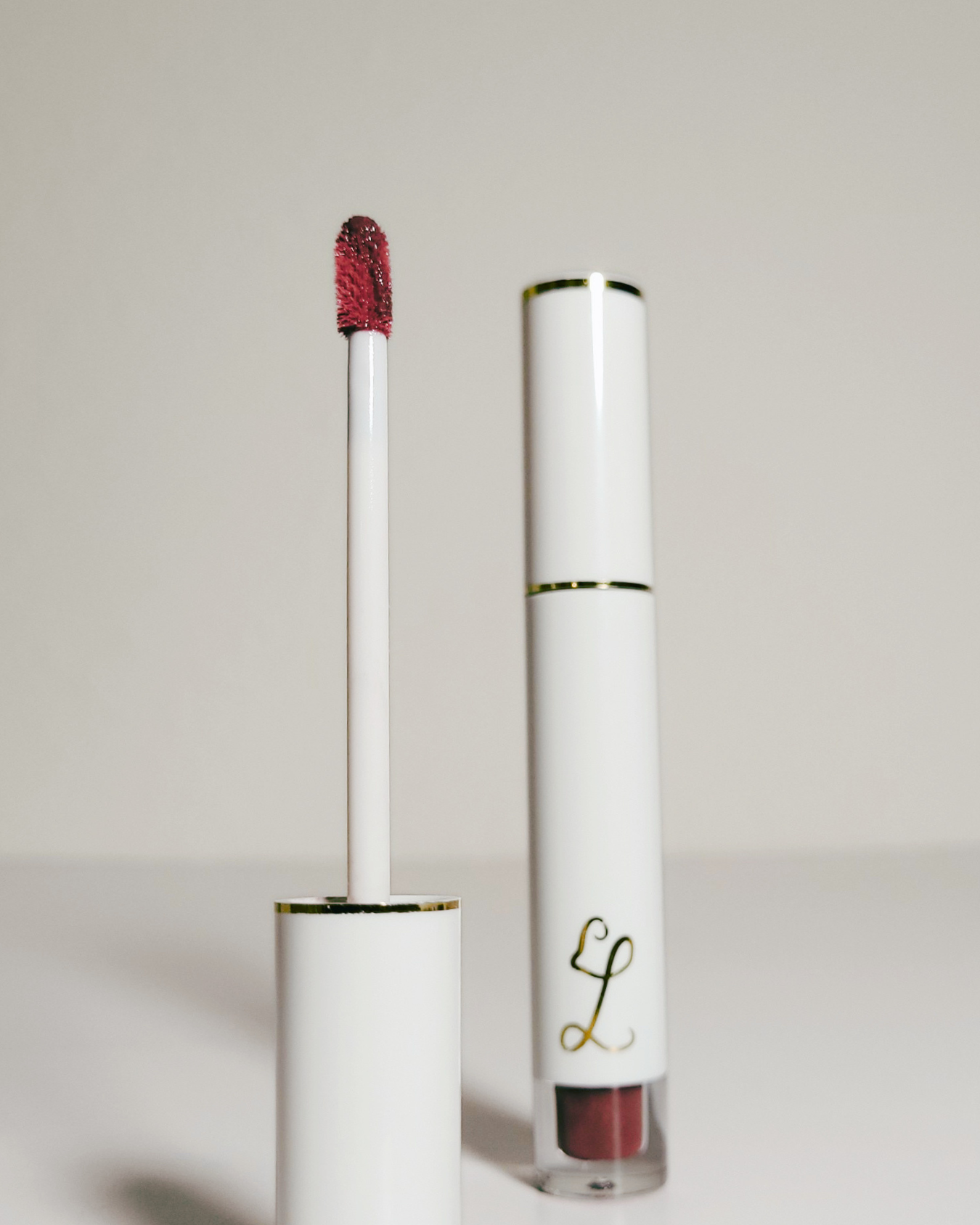 A white liquid lipstick tube with gold trim stands next to its applicator wand, which is loaded with a deep berry red shade, on a clean light gray background.
