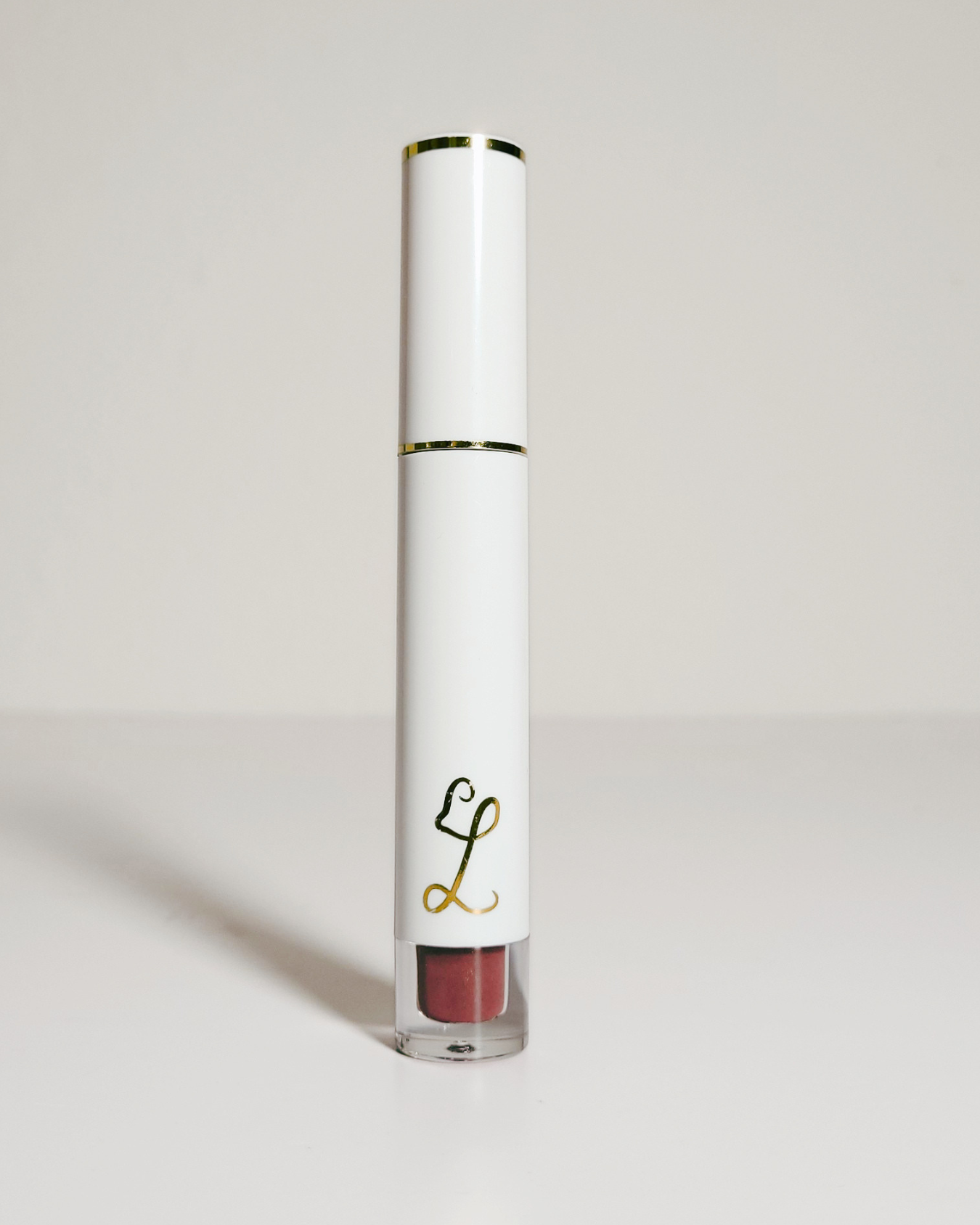 A white cylindrical cosmetic tube with gold trim and a clear base showing a mauve-brown lip product inside, photographed on a clean light gray background.