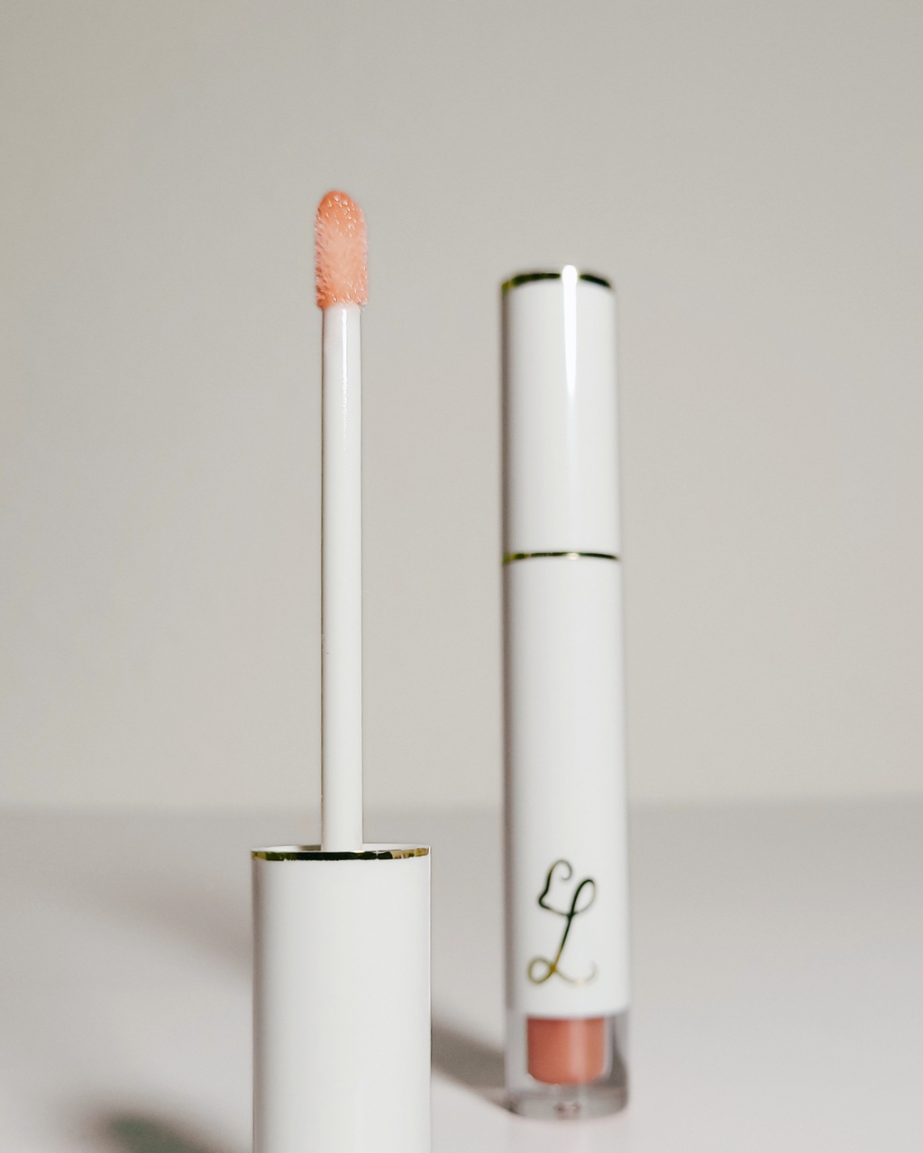 A white cosmetic tube with a gold accent band and a peach-colored applicator wand, shown next to the open base of the tube on a light gray background.