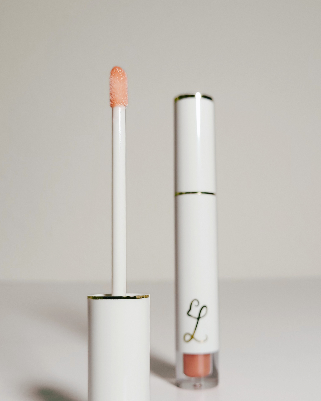 A white cosmetic tube with a gold accent band and a peach-colored applicator wand, shown next to the open base of the tube on a light gray background.