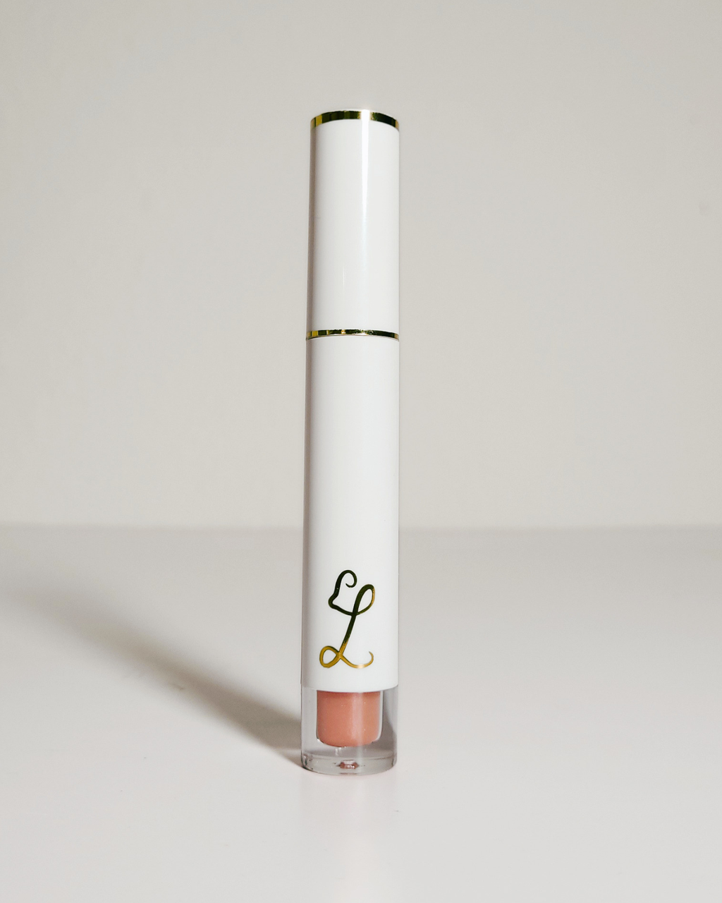 A white cylindrical lip gloss tube with gold trim and a clear base showing a peachy nude gloss, standing on a light gray surface against a light gray background.