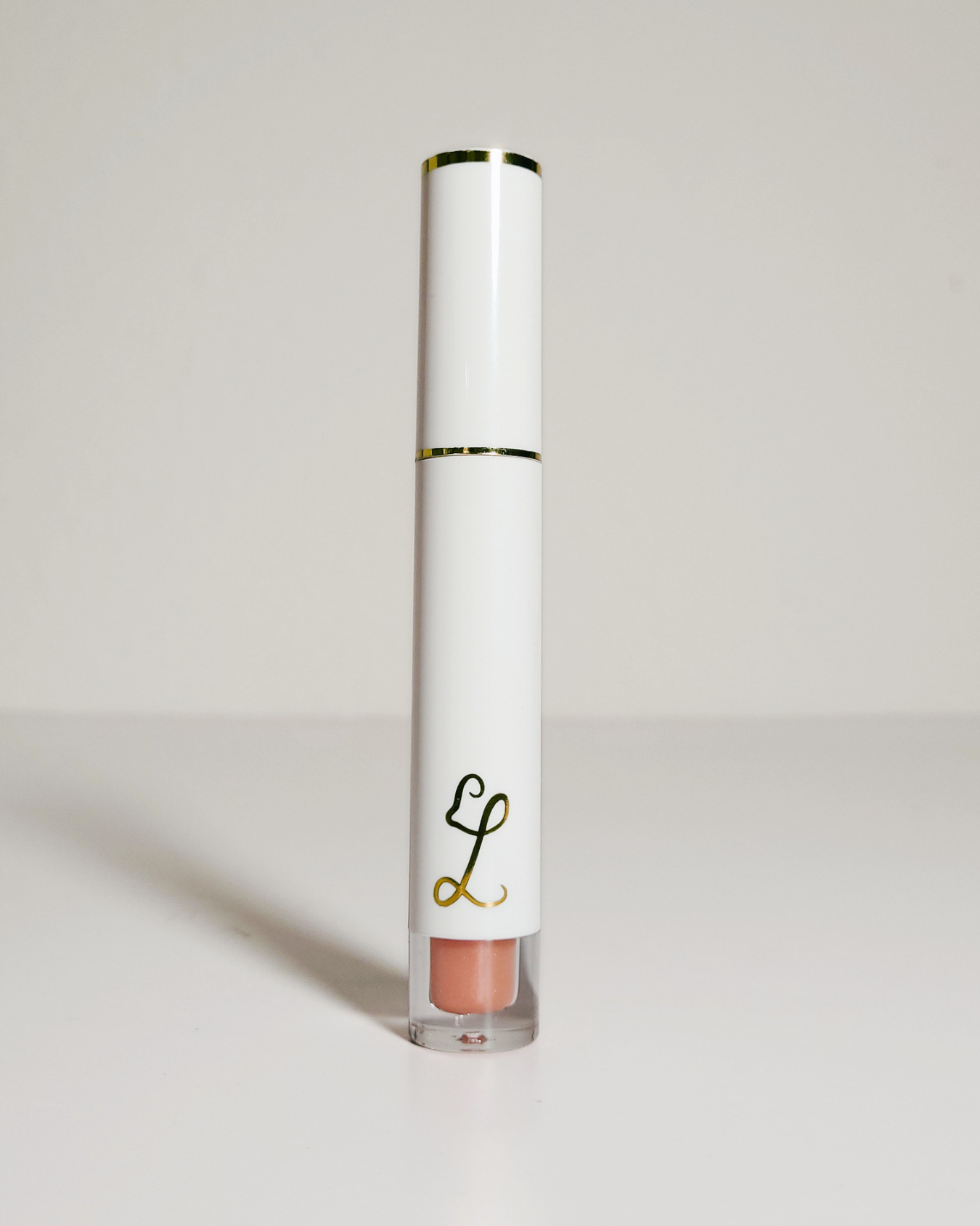 A white cylindrical lip gloss tube with gold trim and a clear base showing a peachy nude gloss, standing on a light gray surface against a light gray background.