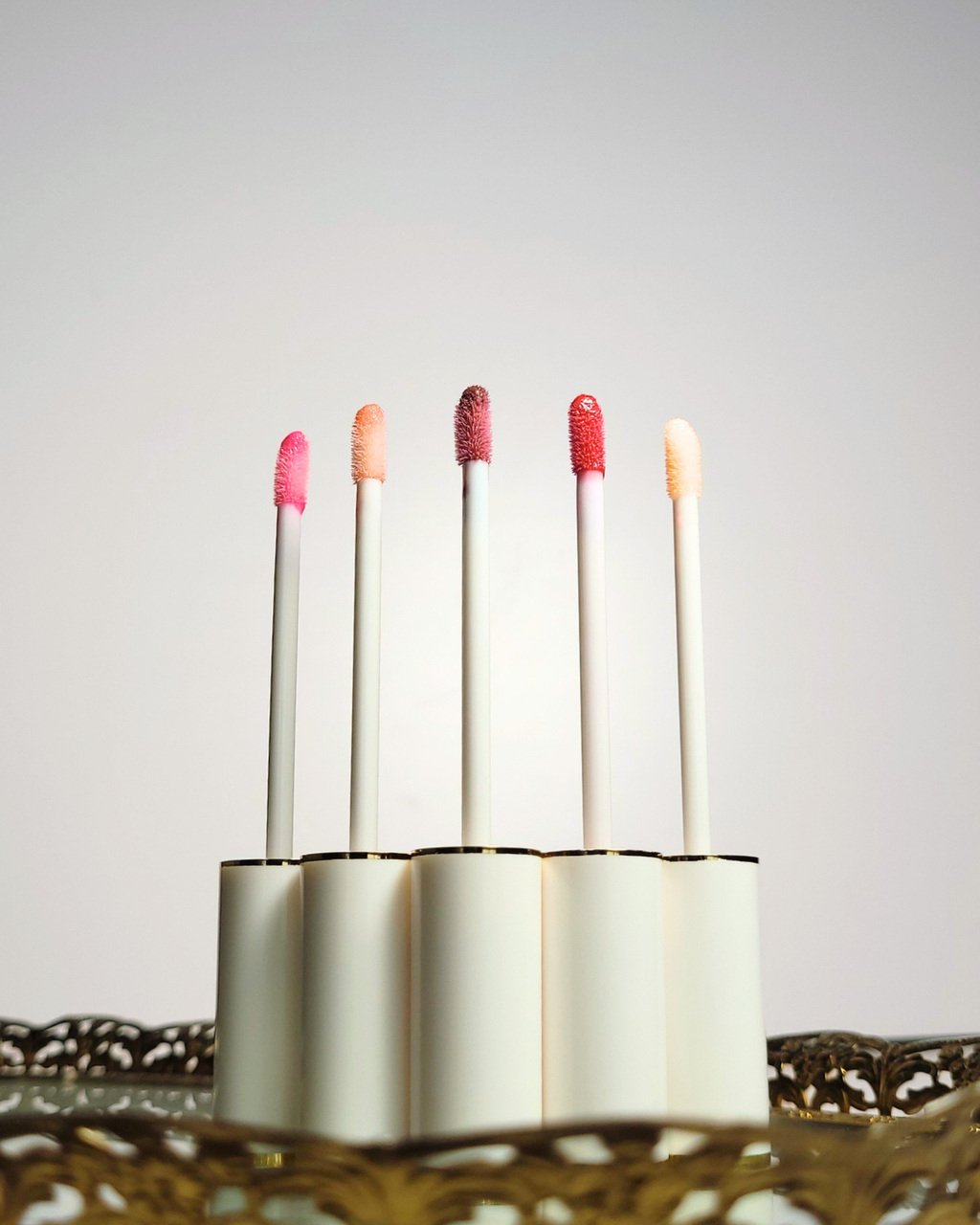 Set of lip glosses with colorful caps in a holder on a light background, Five white and gold lip gloss tubes stand in a row on an ornate vintage gold mirrored tray, showing off clear bases with distinct lip gloss shades ranging from clear pink to deep mauve.