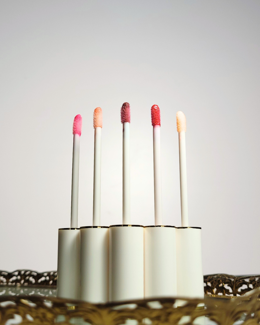 Set of lip glosses with colorful caps in a holder on a light background, Five white and gold lip gloss tubes stand in a row on an ornate vintage gold mirrored tray, showing off clear bases with distinct lip gloss shades ranging from clear pink to deep mauve.
