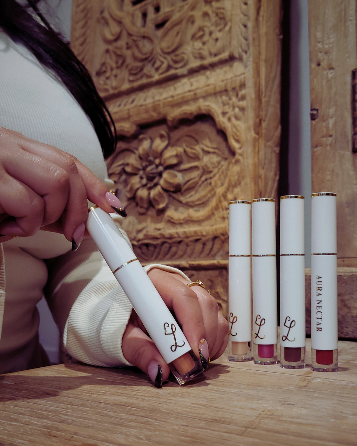 Person holding a white lipstick with Aura Nectar next to four other lipsticks on a wooden surface.