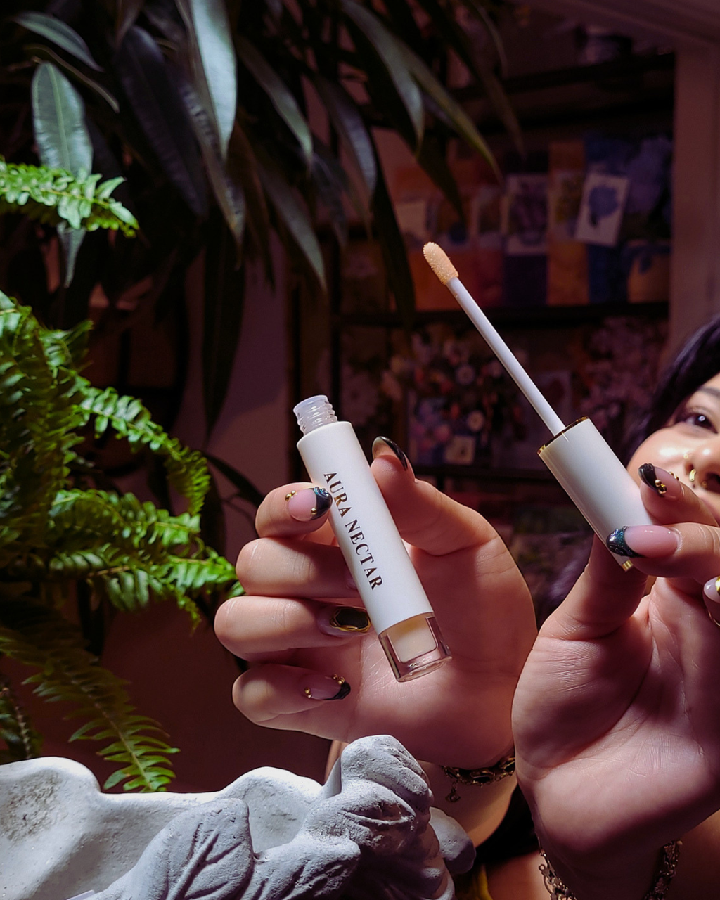 A person holds an open tube of ARJUNTEIR liquid concealer next to its applicator wand, showcasing the product in a lifestyle setting with a dark background and green plants.