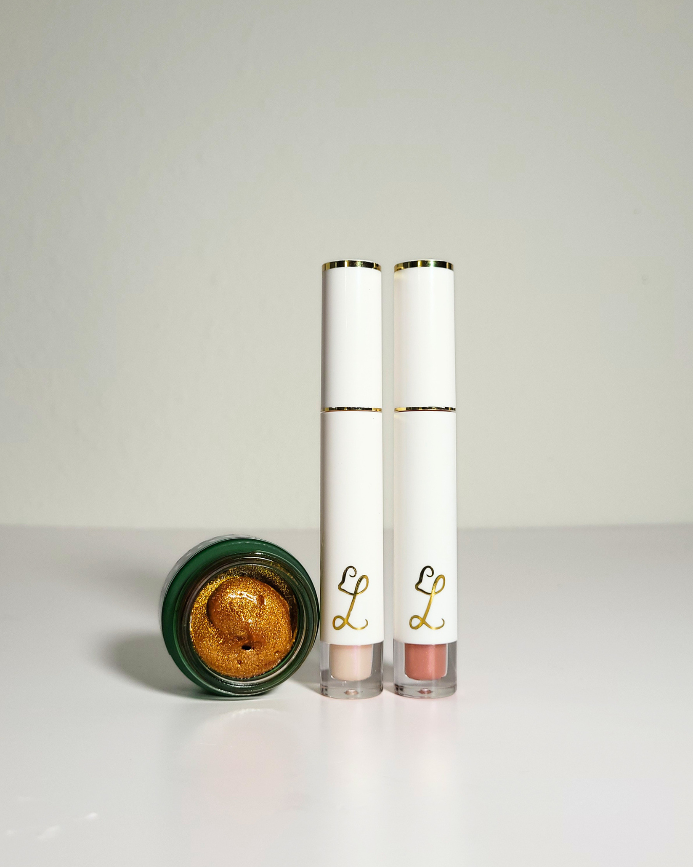 Two white cosmetic tubes with gold lettering and a green-lidded pot of shimmering gold cream, displayed on a light gray background.