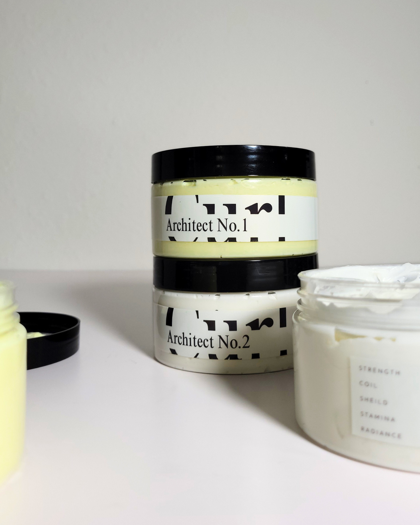 Two white cosmetic jars with black lids on a white surface against a light gray background. One jar is stacked on top of the other, showing the label 'Architect No.