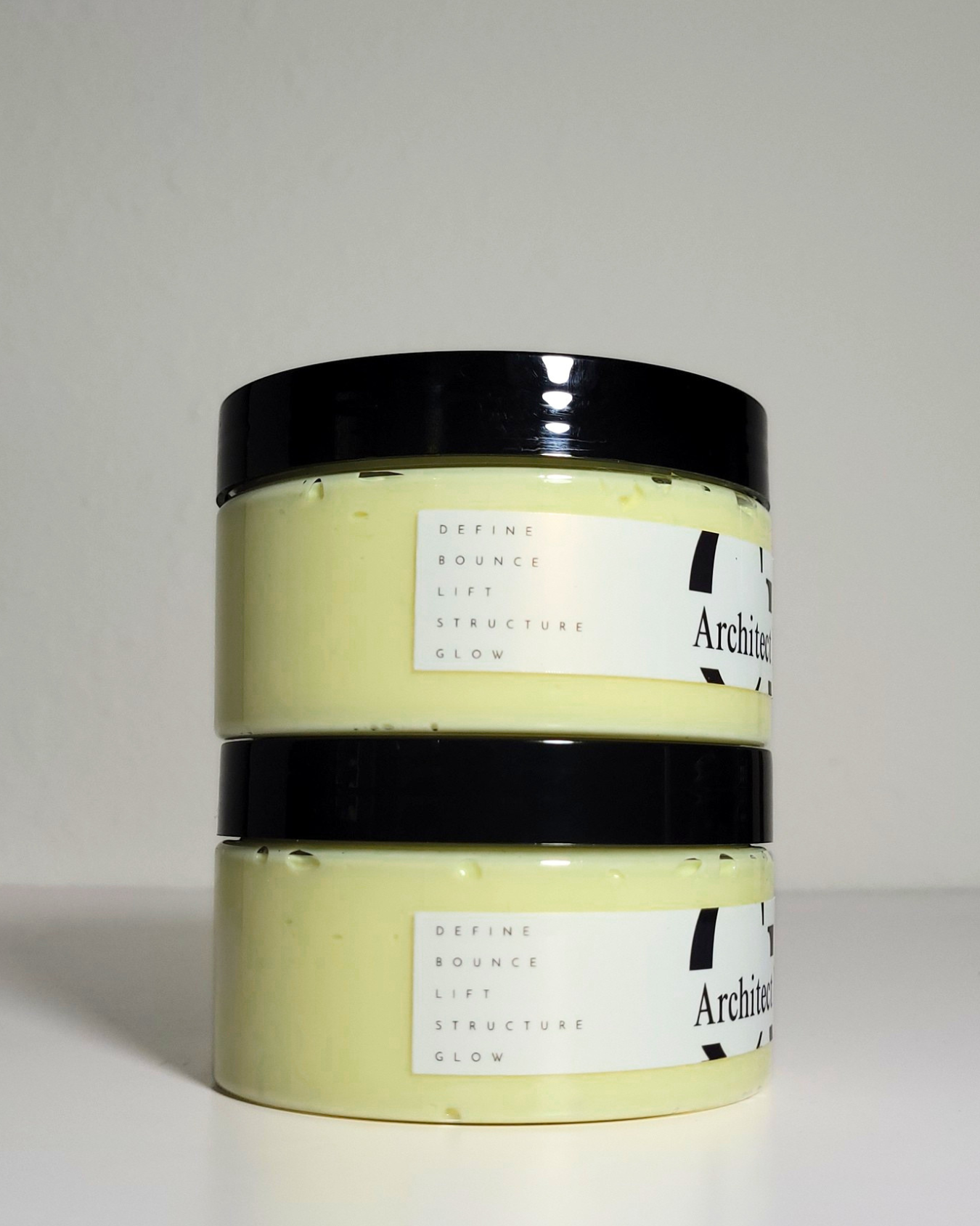 Two stacked jars of Architecte Cosmetics 'Define Bounce Lift Structure Glow' cream in a pale yellow shade, featuring black lids and white labels, set against a neutral light gray background.
