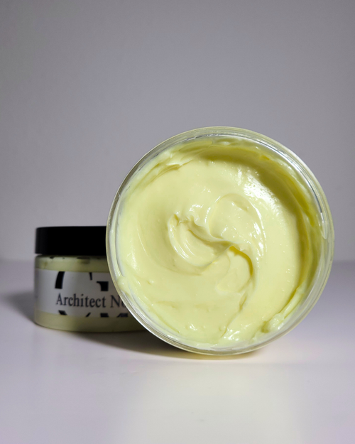 An open jar of pale yellow cream or balm in the foreground with its lid placed behind it on a plain light gray background. The label partially shows the text 'Architect N.