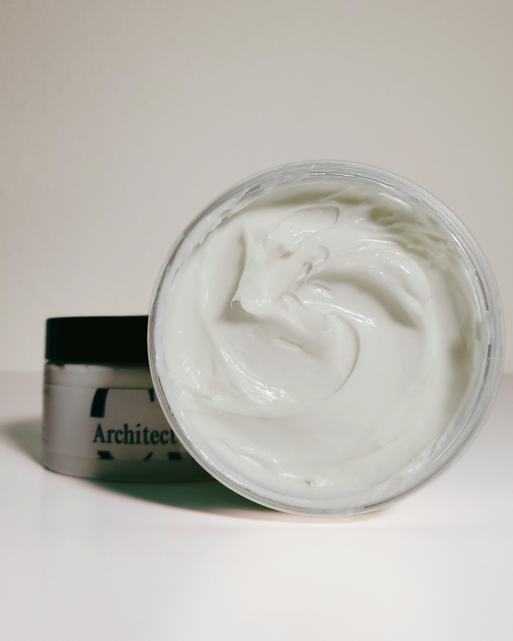 An open clear plastic jar filled with thick white cream, with a silver cylindrical container and black lid standing behind it on a plain light background.