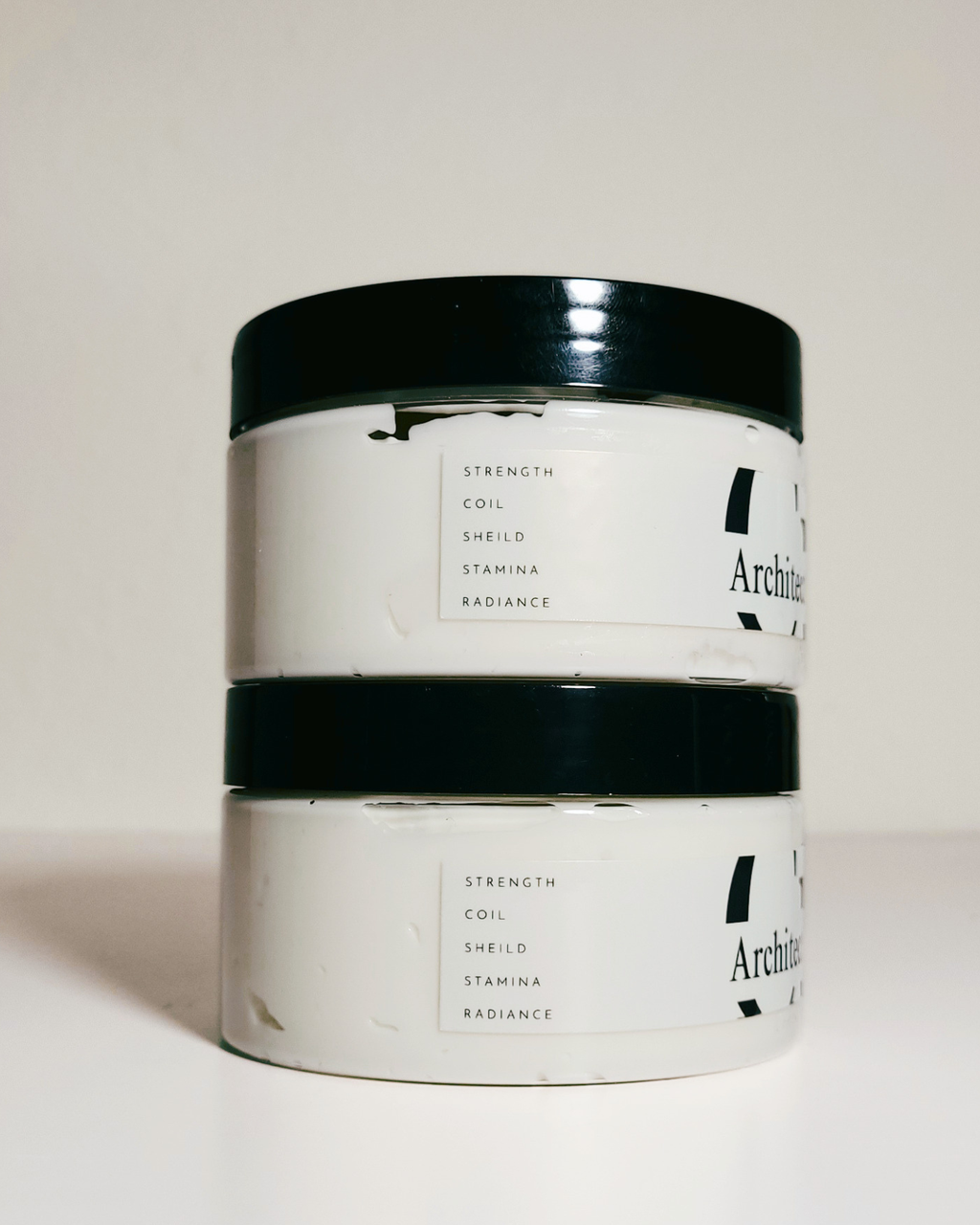Two stacked white plastic jars with black lids, featuring minimalist black text labels on a clean, light beige background.