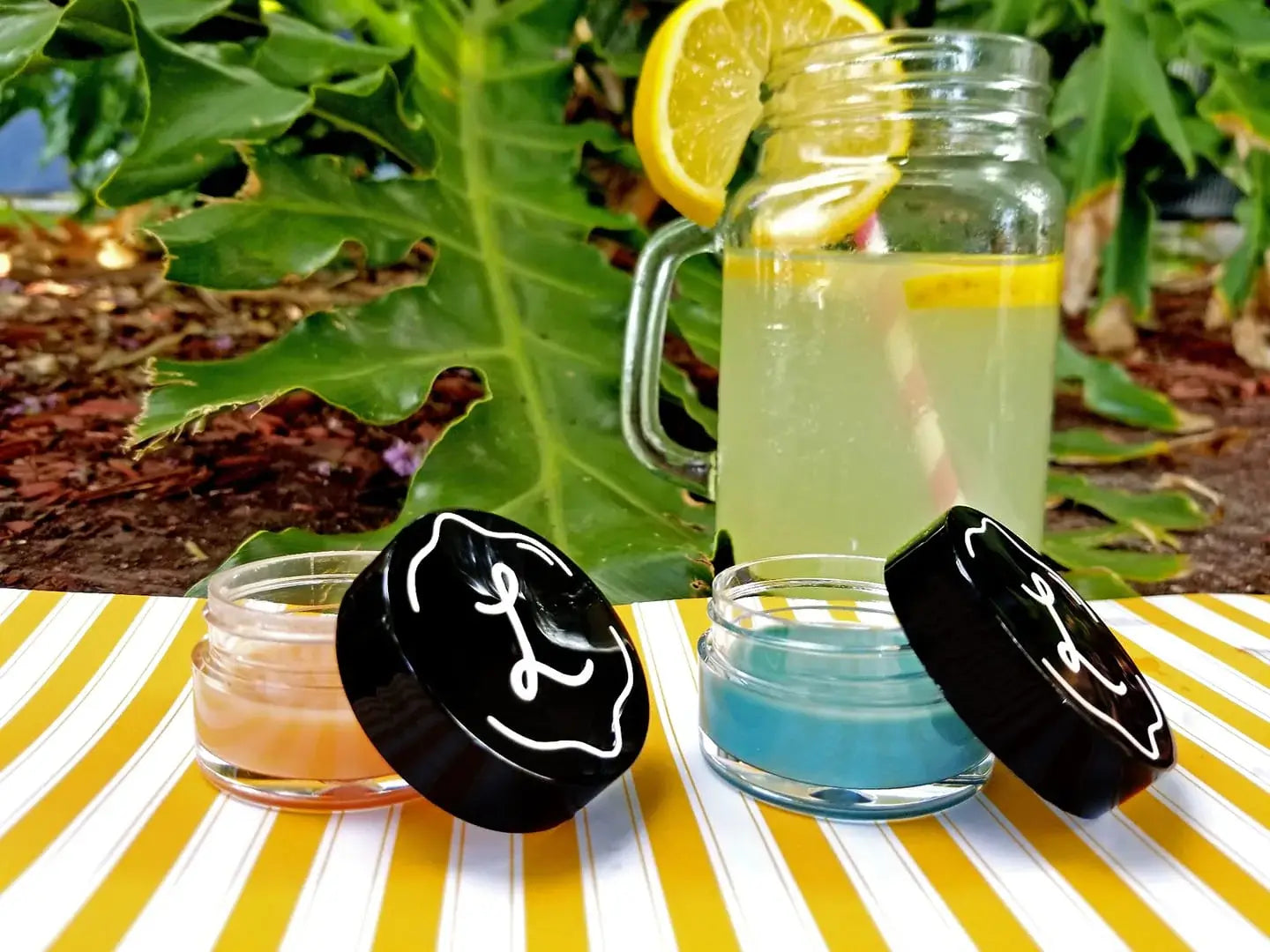 Three glass jars with colorful liquids on a striped surface with greenery in the background with lip balms