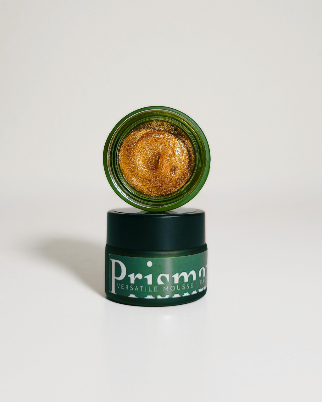 An open jar of Prisma Cascate Mousse Lip in a shimmering gold shade, with the lid propped open above the dark green container on a clean white background.