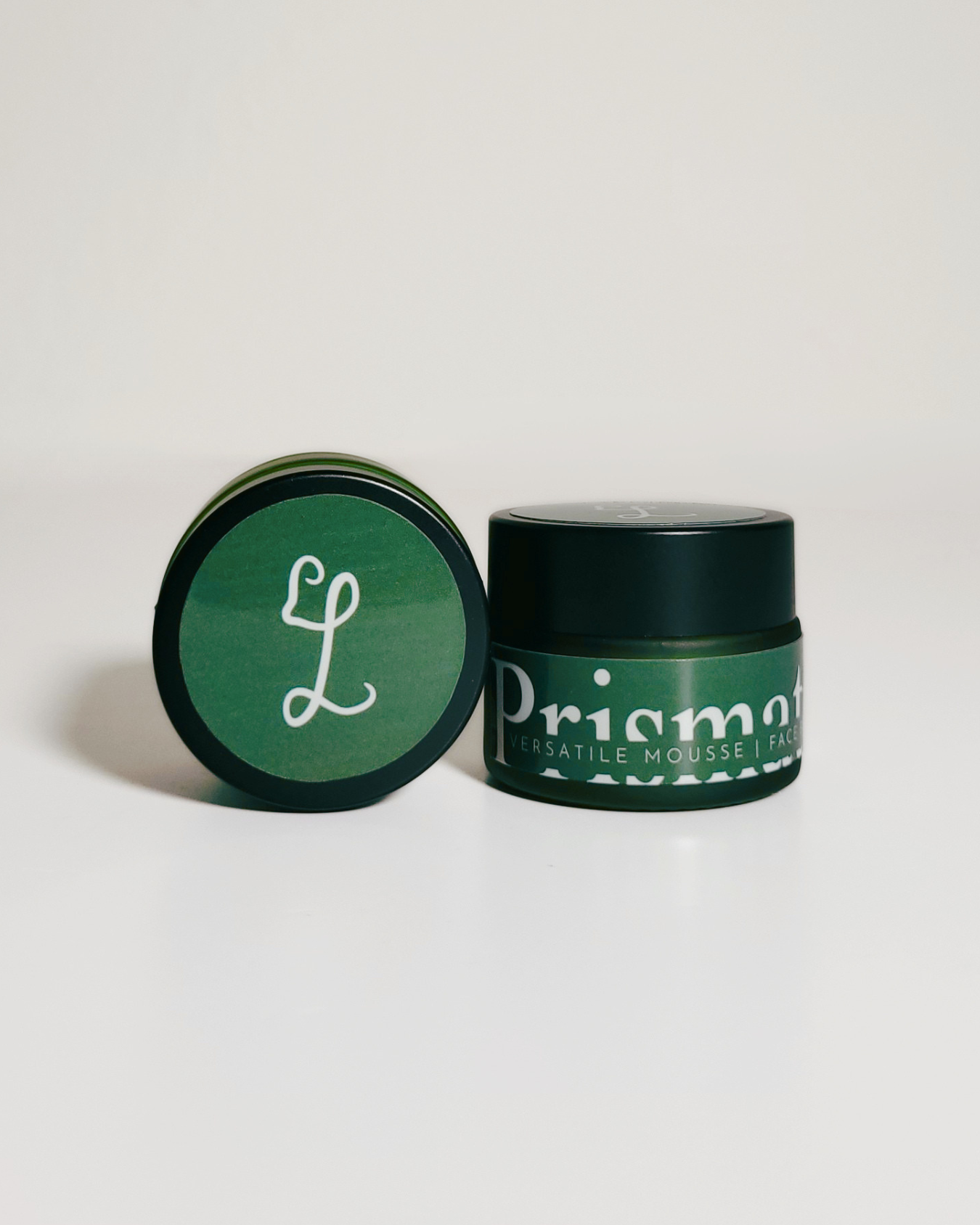 A dark green jar of Priama Cosmétique matte mousse hair wax next to its matching lid, which features a white stylized 'L' logo, on a clean white background.