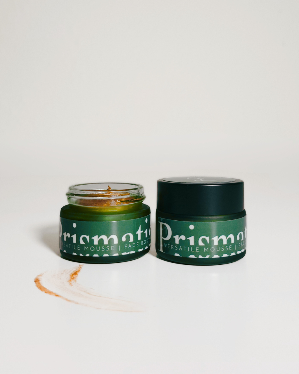 Two green cosmetic jars from Priama's Herbale Mousse Factory on a clean white background, with one jar open showing the product and one closed. A small smear of the product is visible in front.